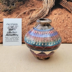 May include: A small, round Native American pottery vase with a narrow neck. The vase features a colorful design with bands of red, blue, and green. A certificate of authenticity is visible in the background.