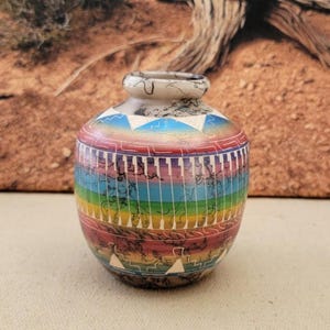 May include: A small, round, hand-painted vase with a rainbow design. The vase features a white rim and a base with a dark, textured pattern. The body of the vase is decorated with horizontal bands of color, including red, blue, and yellow.
