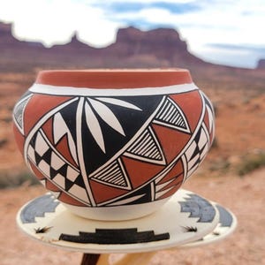 May include: A handcrafted, round ceramic pot with a red-brown base, adorned with black, white, and geometric designs. The pot sits on a decorative stand, with a desert landscape in the background. The pot is likely used for decorative purposes.