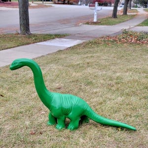 May include: A bright green dinosaur statue with a long neck and tail stands on a grassy lawn. The dinosaur has a textured surface and is positioned outdoors. The background includes a sidewalk, street, and trees.