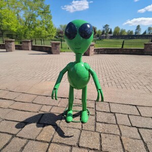May include: A green metal alien sculpture with large black eyes, arms, and legs. The sculpture is standing on a brick surface, with a shadow cast behind it. The alien has a round body and a small smile.