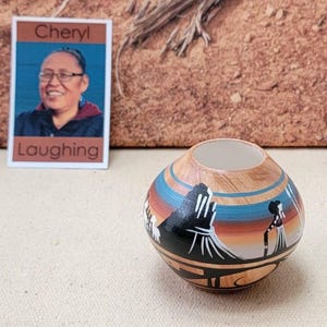 May include: A small, round, hand-painted ceramic vase with a light brown exterior and a white interior. The vase features a design with black, blue, and orange elements. A small card with the text "Cheryl Laughing" is also in the image.