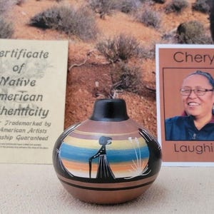 May include: A Native American pottery vase with a black top and a brown base. The vase features a painted scene with a figure and landscape. Includes a certificate of authenticity and a card with the name "Cheryl" and the word "Laughing".