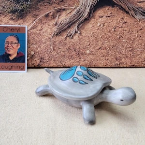 May include: A ceramic turtle figurine with a light gray body and turquoise accents on its shell. The turtle has four legs and a small head. The figurine is on a cream-colored surface. A small card with the text "Cheryl Laughing" is also present.