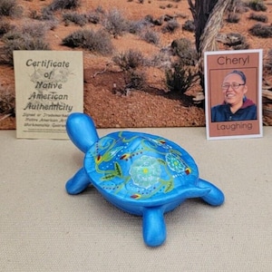May include: A blue ceramic turtle figurine with a detailed, colorful design on its shell. The turtle is displayed with a certificate of Native American Authenticity and a photo of a person named Cheryl.