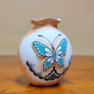 May include: A small, ceramic vase with a butterfly design. The vase is primarily white with a light brown rim and base. A large butterfly with turquoise wings and white accents is centered on the front. Black and turquoise decorative elements are below the butterfly.