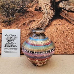 May include: A small, round, Native American pottery vase with a colorful, geometric design. The vase features bands of blue, pink, red, and gold. A certificate of authenticity is visible in the background.