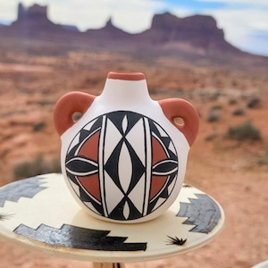 May include: A small, white ceramic vase with two handles and a geometric design in black, red, and white. The vase sits on a round, cream-colored stand with a black and gray border. The background features a desert landscape.