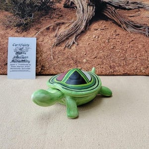 May include: A green ceramic turtle figurine with a geometric design on its shell. The turtle has a light green body and a shell with black, red, and green accents. The figurine is displayed on a cream-colored surface.