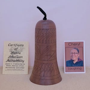 May include: A brown, bell-shaped Native American art piece with carved designs. It has a black cord at the top and is displayed with a certificate of authenticity and a photo of a person named Cheryl. The certificate states it is signed or trademarked by Native American Artists.