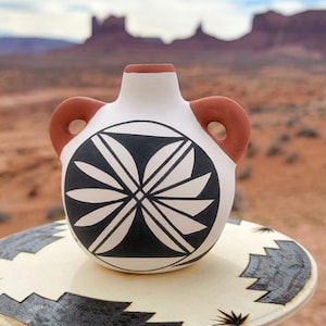 May include: A small, white ceramic vase with a black and white geometric design and two brown handles. The vase is displayed on a decorative stand with a similar pattern. The background features a desert landscape with red rock formations.