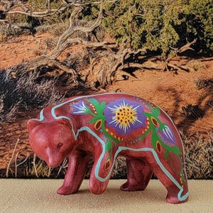May include: A maroon-colored bear figurine with floral designs in green, purple, and yellow. The bear is standing on all fours, with the floral pattern adorning its back and sides. The background features a desert landscape.