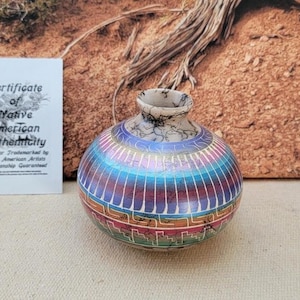May include: A handcrafted, round Native American pottery vase with a colorful, iridescent finish. The vase features a striped pattern in shades of blue, purple, and red, with a geometric design at the base. A certificate of authenticity is included.
