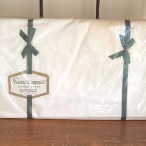 May include: A white Wamsutta Supercale fitted sheet in clear plastic packaging with dark green ribbon ties. A tan label reads Wamsutta Supercale Fitted Sheet for 39 inch by 75 inch mattress. The sheet is folded into a rectangular shape.