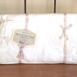 Könnte beinhalten: Ein weißes Spannbettlaken, verpackt in durchsichtiger Folie mit beigen Schleifenbändern. Ein Etikett auf der Vorderseite zeigt die Aufschrift No-Iron Percale, Supercale Plus, 200 Thread Count und Wamsutta. Die rechteckige Packung ist mit Markenband fixiert.