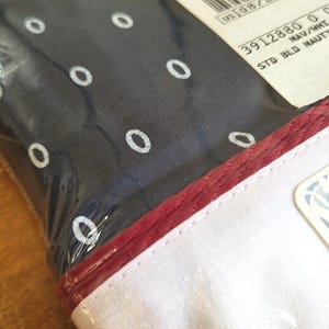 May include: A navy blue fabric with white oval patterns, packaged in clear plastic. The fabric has a red border and a white edge. A tag with blue floral patterns is visible.