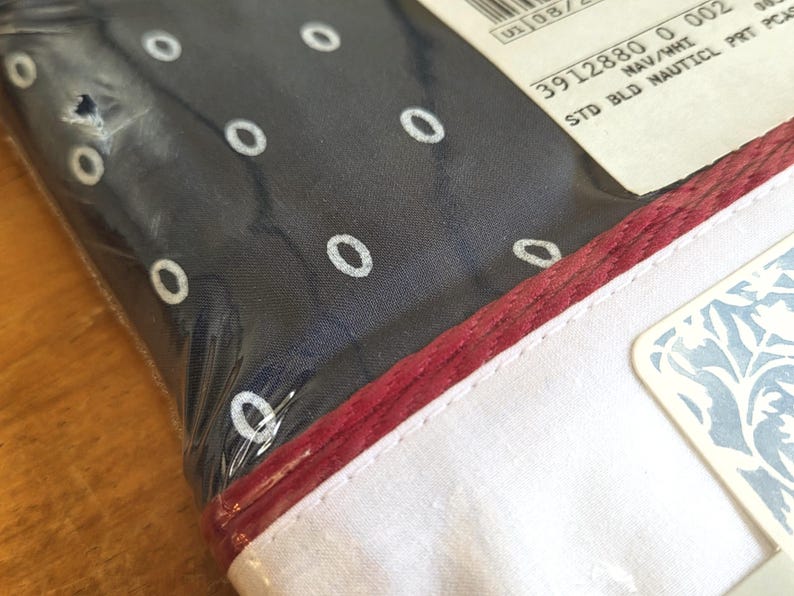 May include: A navy blue fabric with white oval patterns, packaged in clear plastic. The fabric has a red border and a white edge. A tag with blue floral patterns is visible.