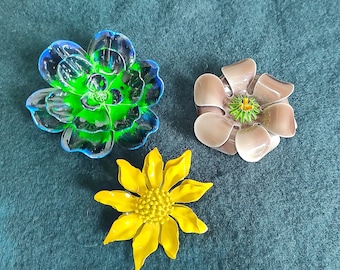 Peut inclure: Trois broches en forme de fleurs. L'une est une fleur transparente, verte et bleue. Une autre est une fleur beige avec un centre vert et jaune. La troisième est une fleur jaune avec un centre texturé.