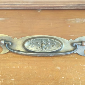 Pode incluir: Puxador de gaveta de latão antigo com um centro oval apresentando uma águia e duas figuras laterais. O puxador tem extremidades curvas e uma alça curva. A ferragem está sobre uma superfície de madeira.