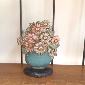 May include: A cast iron doorstop featuring a bouquet of pink and white flowers with green leaves in a teal vase. The base is a solid black semi-circle. The metal surface shows a weathered, textured finish with visible signs of age and wear.