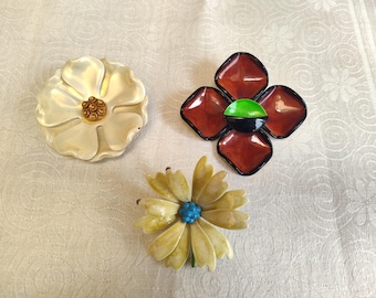 May include: Three vintage flower brooches. One is white with a gold center, one is red with a green and black center, and one is yellow with a blue center.