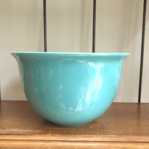 May include: A solid turquoise mixing bowl with a smooth, glossy finish and a wide, flared rim. The bowl features a deep, rounded basin and a consistent, vibrant color throughout its exterior surface, presenting a simple and functional design.