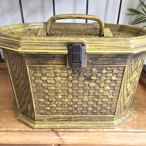 May include: A vintage, octagonal storage box with a woven basket design. The box is yellow with brown accents, a handle, and a clasp closure. The box is made of a hard material and sits on a wooden surface.