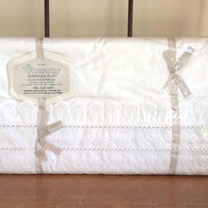 May include: A white Wamsutta Supercale Plus twin flat sheet in original plastic packaging with a beige ribbon. The sheet features a decorative eyelet lace trim along the top edge. The label indicates a finished size of 72 by 96 inches.