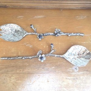May include: A pair of silver-toned metal salad servers featuring handles shaped like textured tree branches. Each handle is adorned with small metal flowers and a single dark purple stone accent. The serving ends are shaped like large leaves.
