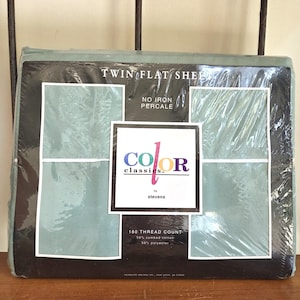 May include: A sealed package of twin flat sheets in a muted teal color. The packaging features the text "TWIN FLAT SHEET" and "NO IRON PERCALE". A central label reads "COLOR classics" by stevens, with a thread count of 180.