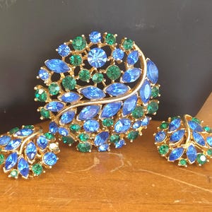 May include: A vintage jewelry set featuring a large brooch and matching clip-on earrings. The set has a gold-tone metal base and is adorned with blue and green rhinestones. The brooch and earrings have a leaf-like design.