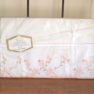 May include: A white sheet set in clear plastic packaging featuring a delicate embroidered pattern of pink flowers with green stems along the bottom edge. A hexagonal label reads Wamsutta Supercale Plus Sheets and Pillowcases 90 x 102.