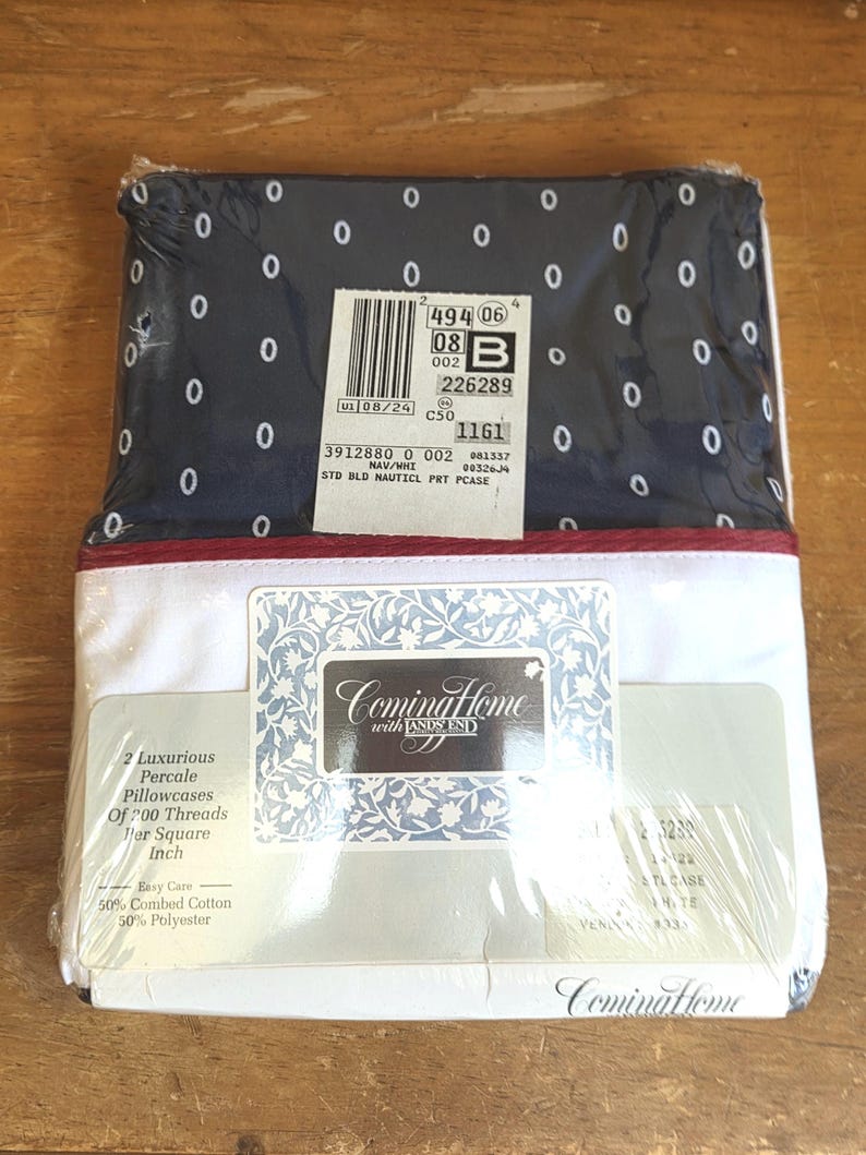May include: A packaged set of Coming Home brand pillowcases. The top of the package is navy blue with white oval patterns. The bottom is white with a red stripe. The package includes two percale pillowcases made of 50% combed cotton and 50% polyester.