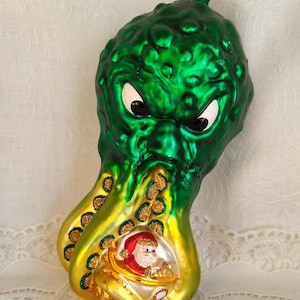 May include: Green glass Christmas ornament shaped like a monster with a Santa Claus inside. The monster has large eyes and a textured surface.