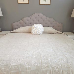 May include: A bed with a tufted headboard, a cream-colored floral patterned quilt, and a decorative white pillow. Two framed floral prints hang above the bed. The bed is flanked by two white nightstands with lamps.
