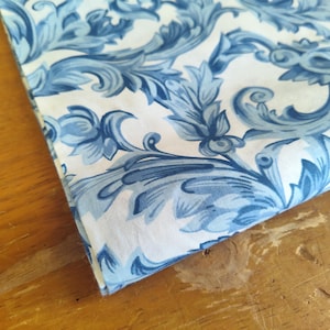 K&ouml;nnte beinhalten: Wei&szlig;er Stoff mit einem blauen und wei&szlig;en Blumenmuster. Der Stoff hat ein Damastdesign mit komplizierten Wirbeln und Bl&auml;ttern.