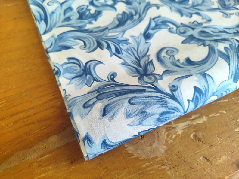 K&ouml;nnte beinhalten: Wei&szlig;er Stoff mit einem blauen und wei&szlig;en Blumenmuster. Der Stoff hat ein Damastdesign mit komplizierten Wirbeln und Bl&auml;ttern.