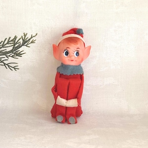 May include: A vintage elf doll with a red outfit, hat, and shoes. The doll has a painted face with blue eyes, rosy cheeks, and a small smile. A gray collar and white belt complete the festive look. The doll is approximately 6 inches tall.