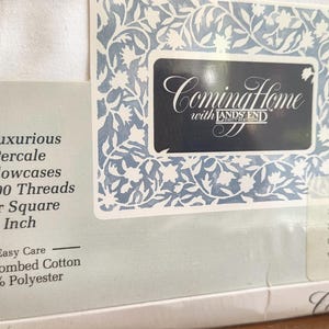 May include: White percale pillowcases from Lands' End, featuring a floral design and the text "Coming Home." The packaging indicates 200 threads per square inch, 50% combed cotton, and 50% polyester. The label has a blue and white floral border.
