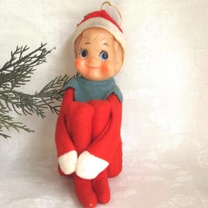 Puede incluir: Una muñeca elfo de estilo vintage con cara de plástico moldeado, mejillas sonrosadas y ojos azules. La muñeca viste un traje de fieltro rojo con manoplas blancas, un cuello azul y un gorro rojo con un borde blanco.