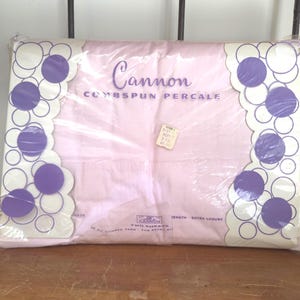 May include: A package of Cannon Combspun Percale sheets in a light pink color. The packaging features a white border with purple circles and the text Cannon Combspun Percale. A small price tag is attached to the center of the plastic wrapping.