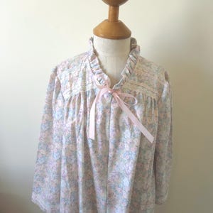 May include: A floral-print robe in pastel pink, blue, and lavender. The robe features a ruffled collar, lace trim, and a pink ribbon tie. The long-sleeved robe is displayed on a mannequin.