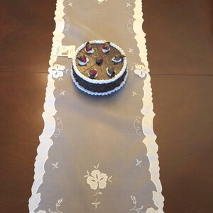 Könnte beinhalten: Weißer, bestickter Tischläufer mit gewelltem Rand und floralen Mustern. Ein Schokoladenkuchen mit weißer Glasur und Fruchtbelag liegt darauf. Der Tischläufer ist etwa 152 cm lang.