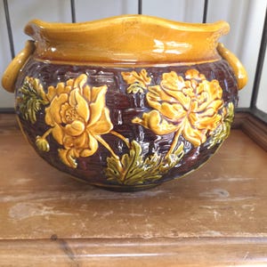 May include: A round ceramic planter featuring a glossy mustard yellow rim and handles, with a dark brown basket-weave textured body. The exterior is decorated with raised, relief-style yellow flowers and green leaves wrapping around the center.