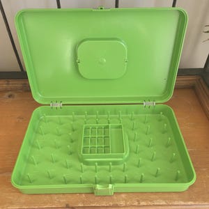 May include: Open, bright green plastic thread organiser with a hinged lid. The interior features numerous pegs for holding spools of thread, along with a small compartment. The organiser is designed to keep threads neat and accessible.