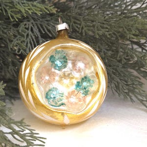 May include: A vintage Christmas ornament with a gold and white design. The glass orb features a clear center with teal and pink floral accents. The ornament is suspended from a silver metal cap and is set against a backdrop of evergreen branches.