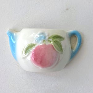 Può includere: Un vaso da parete in ceramica bianca a forma di teiera in miniatura. La parte anteriore presenta un disegno dipinto a mano di una pesca rosa con foglie verdi e un piccolo fiore blu. Il beccuccio e il manico sono smaltati in azzurro.
