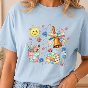 May include: A light blue t-shirt featuring a colorful graphic print of a yellow smiley face, a brass hand bell with a plaid bow, a cup filled with colored pencils, a stack of books, and small floral and star accents scattered throughout.