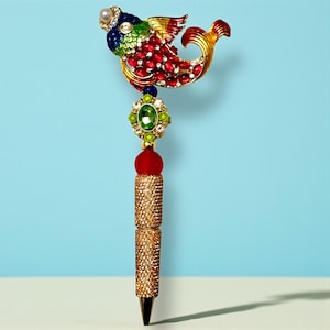 May include: A decorative pen featuring a bejeweled fish design in red, gold, blue, and green. The pen's body is gold with a textured pattern, and it has a black tip. The fish has a pearl at the top and is adorned with red and clear gemstones.