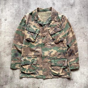 Könnte beinhalten: Tarnmusterjacke in Grün-, Braun- und Beigetönen. Sie hat vier Vordertaschen und einen Kragen. Der Stoff des Kleidungsstücks weist Gebrauchsspuren auf, was ihm eine Vintage- oder Militärästhetik verleiht.